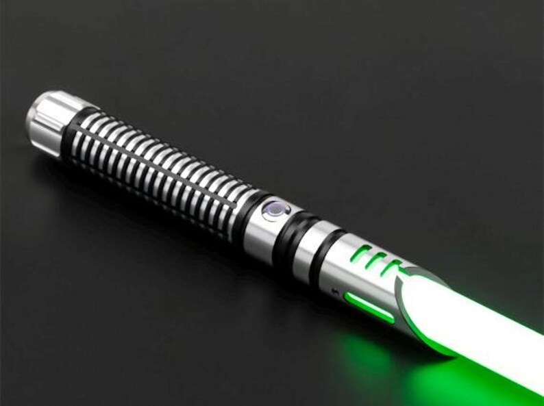 Pu&ograve; includere: Una spada laser argentata e nera con una lama verde brillante. L'elsa ha un design a coste e un pulsante. La lama si illumina intensamente, suggerendo che sia attivata. La spada laser &egrave; ambientata su uno sfondo scuro.