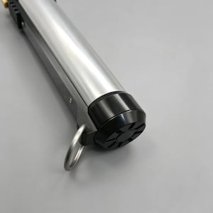 May include: A silver and black cylindrical object with a ring attached. The object has a metallic silver body and a black base with a starburst pattern. The object is likely a tool or a part of a larger device.