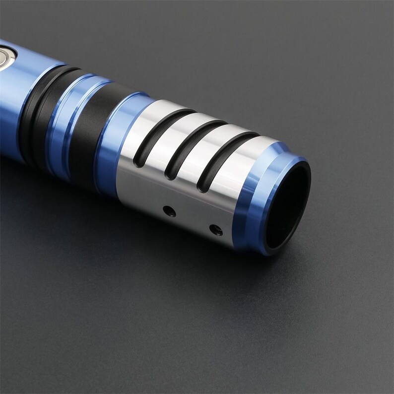 May include: Close-up of a blue and silver lightsaber hilt. The hilt features a silver section with four angled cutouts, a black band, and blue accents. The end of the hilt is a dark, open cylinder.