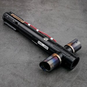 K&ouml;nnte beinhalten: Ein schwarzes Lichtschwert mit einem Kreuzguard-Design. Der Griff hat silberne und goldene Akzente, mit einem roten Kabel. Die Enden des Kreuzschutzes haben ein metallisches, irisierendes Finish.