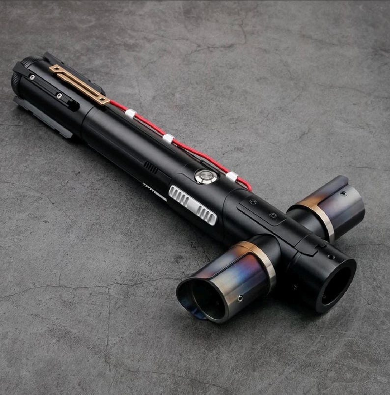 K&ouml;nnte beinhalten: Ein schwarzes Lichtschwert mit einem Kreuzguard-Design. Der Griff hat silberne und goldene Akzente, mit einem roten Kabel. Die Enden des Kreuzschutzes haben ein metallisches, irisierendes Finish.