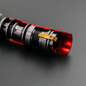 May include: Close-up of a red and silver lightsaber hilt. The hilt features a black grip, silver accents, and a gold-colored clamp. The lightsaber is partially cut away, revealing internal components.