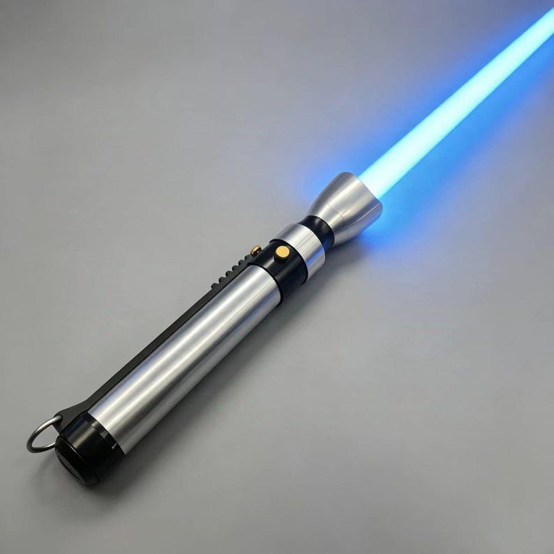 May include: A silver and black lightsaber with a bright blue blade. The hilt features a metallic finish with black accents, a gold button, and a ring at the base. The glowing blue blade extends from the top.