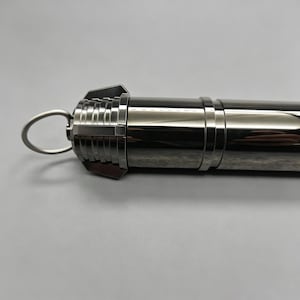 Op de afbeelding: Een metallic zilveren cilindrisch object met een lus aan &eacute;&eacute;n uiteinde. Het object heeft een reeks ribbels en een ring in het midden. Het algehele ontwerp suggereert een strakke, moderne esthetiek.