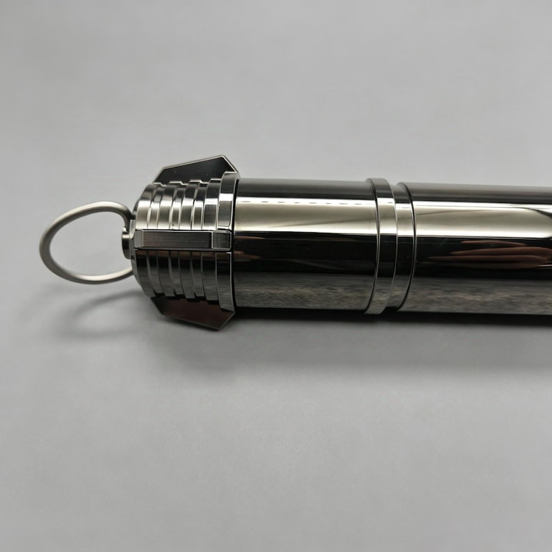 Op de afbeelding: Een metallic zilveren cilindrisch object met een lus aan &eacute;&eacute;n uiteinde. Het object heeft een reeks ribbels en een ring in het midden. Het algehele ontwerp suggereert een strakke, moderne esthetiek.