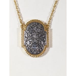 May include: A gold-tone necklace featuring an oval pendant with a scalloped edge. The center of the pendant displays a dark, oxidized silver-tone relief carving of a bird perched amidst intricate floral and leafy botanical patterns.