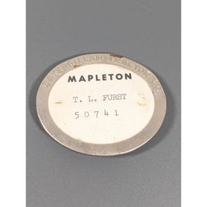 May include: Oval metal Caterpillar Tractor Co. badge with the text "MAPLETON" and "T. L. FURST" printed on a white background. The number "50741" is also printed on the badge. The badge has a silver border.