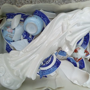 May include: A white porcelain figurine of a woman in a flowing robe, surrounded by broken pieces of blue and white porcelain plates and bowls.