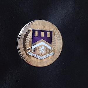 May include: A gold-colored round pin with a purple and white crest featuring a lion and the text "BELLO AC PACE PARATUS".