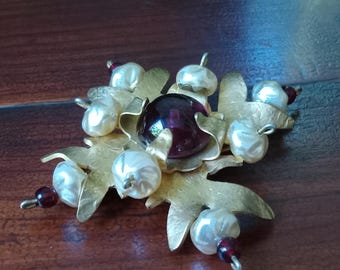 May include: A gold-toned brooch with a large, dark red gemstone in the center. The brooch is decorated with small, white pearls and has a snowflake-like design.