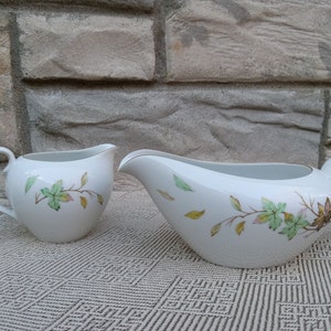 May include: A white porcelain creamer and gravy boat with a floral design. The design features green and brown leaves and branches on a white background.