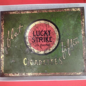 May include: A vintage green and gold Lucky Strike cigarette tin with the words "flat fifties" and "Lucky Strike It's Toasted" printed on it.