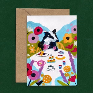 May include: A whimsical greeting card depicting a badger enjoying a tea party. The illustration shows the badger at a table with cakes and tea, set amidst vibrant flowers and a blue and white backdrop. The card is accompanied by a brown envelope.