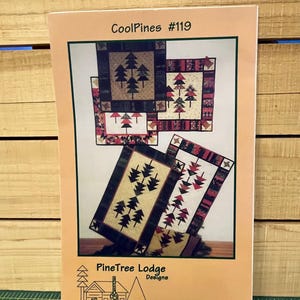 May include: A brochure titled "Cool Pines #119" featuring quilt patterns with pine tree designs. The patterns include various sizes and color schemes, with a logo for "PineTree Lodge Designs" at the bottom. The brochure is on a wooden surface.