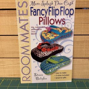 May include: A book cover for "Fancy Flip Flop Pillows" by Donna Babylon. The cover features three decorative flip-flop pillows with floral, leopard print, and striped designs. Text includes "More Splash Than Cash" and "Roommates."