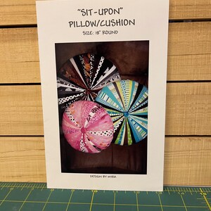 May include: A white card displays a photo of three round pillows/cushions. The pillows are patterned in black and white, pink, and blue. Text on the card reads "SIT-UPON" PILLOW/CUSHION, SIZE: 18" ROUND, and DESIGN BY MYRA.