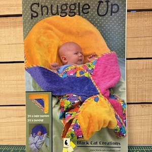 May include: A baby swaddled in a colorful blanket, nestled in a plush orange and pink bunting. The image features the text "Snuggle Up" and "Black Cat Creations". The blanket has a vibrant pattern with purple, green, and blue accents.