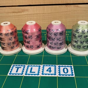 May include: Four spools of thread in shades of pink, peach, lavender, and green. Each spool has a white label with a repeating pattern of a whale.