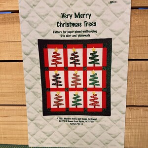 May include: A pattern for a Christmas-themed quilt. The design features nine Christmas trees in red and green, each topped with a gold star. The text reads "Very Merry Christmas Trees" and includes instructions for a wallhanging, tree skirt, and placemats.