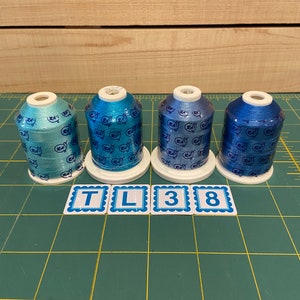 May include: Four spools of blue and teal embroidery thread with a metallic sheen. The spools are labeled "TL38" on a green cutting mat.