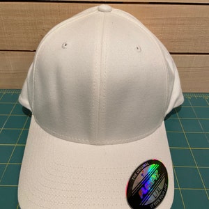 May include: White baseball cap with a Flexfit logo on the front. The logo is a silver circle with black text that reads "The Original Flexfit 1/1 L/XL It Fits Your Life".