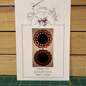 May include: A white paper with the text "Lily Anna Stitches" and a floral illustration. The paper features two circular designs resembling candy corn and summer slices. The text "Candy Corn & Summer Slices Pattern LAS200" is also present.