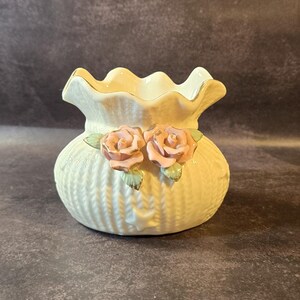 May include: A small, off-white ceramic vase with a ruffled rim and a textured, basket-weave design. Two pink roses are affixed to the front. The vase is a decorative item, suitable for small flowers or as a standalone accent piece.