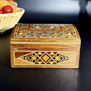 Puede incluir: Una pequeña caja de madera con intrincados patrones geométricos incrustados en tonos marrones, amarillos y negros. La caja está cerrada y se encuentra sobre una superficie negra.