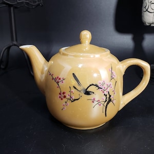 Puede incluir: Una tetera amarilla con un pájaro negro y flores rosas pintadas en ella. La tetera tiene una tapa y un pico.