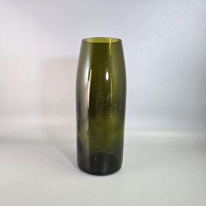 Peut inclure: Un vase cylindrique et élancé en verre vert foncé. Le vase a une ouverture légèrement évasée et une surface lisse et polie. Le vase est vide et posé sur une surface blanche.