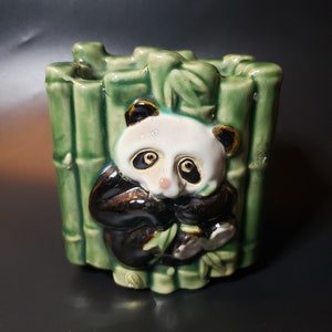 Könnte beinhalten: Ein Keramik-Pflanztopf in Form eines Pandas, der in einem Bambuswald sitzt. Der Topf ist grün und braun mit schwarzen und weißen Details.