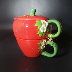 Op de afbeelding: Een tweedelige aardbeivormige theeset van keramiek. De set bevat een theepot met deksel en een bijpassende kop, beide in felrood met witte zaaddetails. Groene handvatten en bloemmotieven dragen bij aan het grillige ontwerp.