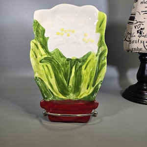 May include: A ceramic poinsettia-shaped dish with a red base and silver-colored metal accents. The dish features a white flower with yellow accents and green leaves. The design is hand-painted and decorative.