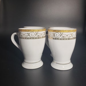 Puede incluir: Cuatro tazas de cerámica blanca con ribete dorado y un diseño floral negro y dorado. Las tazas tienen una base redondeada y un asa.