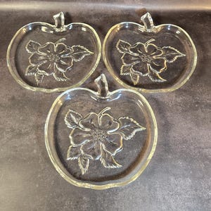 May include: Three clear glass plates shaped like apples, each featuring a molded floral design in the center. The plates have a smooth, transparent finish with a raised stem detail at the top, showcasing a detailed flower with leaves.