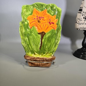 May include: A hand-painted ceramic flower pot with a vibrant orange lily design. The lily has detailed petals and red accents, set against a backdrop of green leaves. The pot is brown and rests on a silver metal stand, creating a decorative piece.