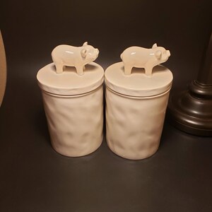 May include: Two off-white ceramic canisters with textured sides and pig-shaped lid handles. The cylindrical canisters sit on a dark surface. The pig figurines are a light cream colour.