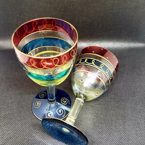 May include: Two hand-painted wine glasses with colorful stripes and gold accents. The glasses are blue, red, yellow, and green.