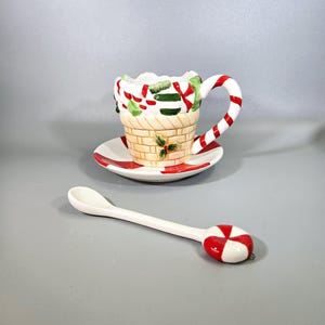 May include: A white ceramic teacup and saucer set with a Christmas candy cane design. The cup has a candy cane handle and the saucer has a red and white candy cane border. The set comes with a white ceramic spoon with a red and white candy cane design on the end.