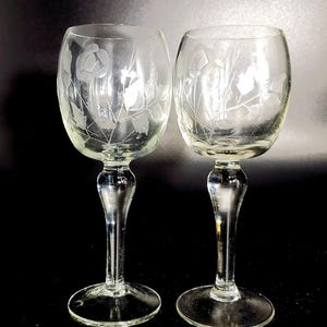 Puede incluir: Dos copas de vino de vidrio transparente con diseños florales grabados. Las copas tienen una copa redonda y un tallo largo y delgado.