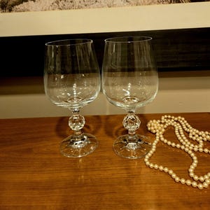 May include: Two clear glass wine glasses with a faceted stem and base. The glasses are on a wooden surface with a string of white pearls.