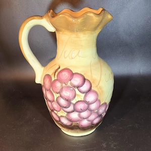 May include: A ceramic pitcher with a brown and yellow glaze and a cluster of pink grapes painted on the side. The pitcher has a scalloped rim and a handle.