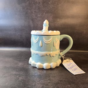 May include: A light blue ceramic mug shaped like a tiered cake, with white frosting details and a candle on top. The mug has a handle and a matching lid. A tag that says "ALL ABOUT YOU" is attached.