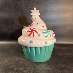 Könnte beinhalten: Ein keramikförmiger Behälter in Cupcake-Form mit sternförmigem Aufsatz. Das Frosting ist rosa mit roten und grünen Süßigkeitenverzierungen. Die Cupcake-Basis ist türkis. Ein dekorativer Artikel.