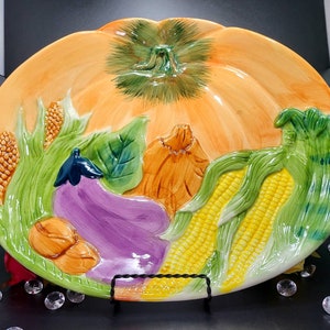 May include: A ceramic serving platter shaped like a pumpkin with a raised design of vegetables, including corn, eggplant, and squash. The platter is painted in shades of orange, green, purple, and yellow.