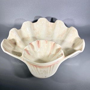 Puede incluir: Un plato de servir de cerámica blanca con forma de concha marina con un tazón más pequeño en el centro. El plato tiene un esmalte rosa y blanco.