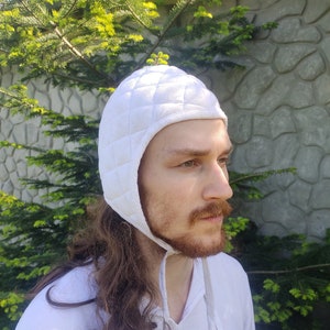 May include: A white quilted coif with a long tie. The coif is worn by a person with long brown hair and a beard.