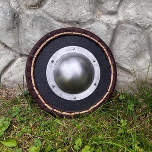 May include: Round shield with a black center, a silver metal boss, and a brown leather border. The edge is trimmed with a rope-like material. The shield is set against a stone wall and green grass.
