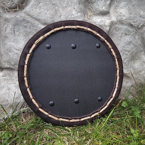 May include: A round, black shield with a brown leather border and a decorative rope trim. The shield has five black rivets and is set against a stone wall and grass.