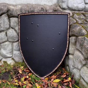 May include: A black shield with a brown border and rivets. The shield is a dark color and has a rounded bottom. The border is a lighter brown color. The shield is leaning against a stone wall.